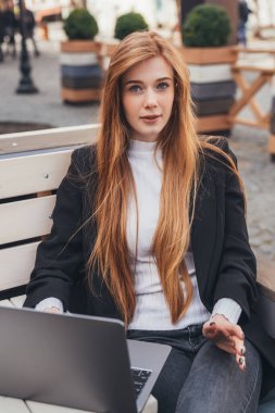 Front view portrait, young freckled woman, resting hands on keyboard, browsing information on computer laptop, sitting in the city park. Education internet