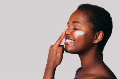 Free space for text. Profile view of an African woman touching her nose with her index finger applying cream on her face, isolated over gray background with