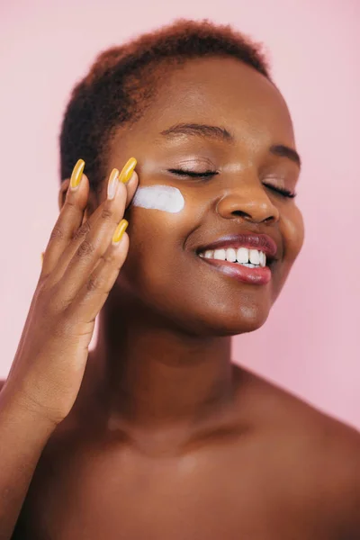 Beautiful african american woman applying moisturizing cream on her cheek, isolated over pink background. Anti-aging skincare. Beauty woman face skin care
