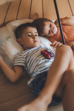 Two children made a hut out of an umbrella and beds, sitting quietly together under the umbrella. Resting, lying asleep, feeling lack of energy.
