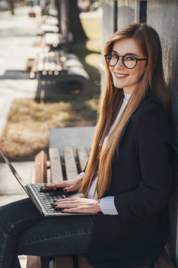 Young ginger student woman preparing for exams with laptop while sitting on wooden bench in the city park. Modern lifestyle. Summer vibes. Outdoor workplace.