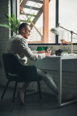 Side view of a man wearing eyeglasses sitting at home listen audio course, checking records at daybook. Studying remotely on quarantine. Homeschooling concept