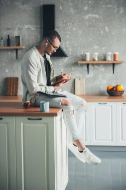 Young man student sitting in the kitchen surfing internet using tablet write up information in his notebook. Learning online. Studying remote training course.