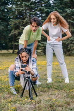 Group of three young teenagers friends wanting to take a group selfie with a selfie stick, standing in city park. Social media trends. Friendship day.