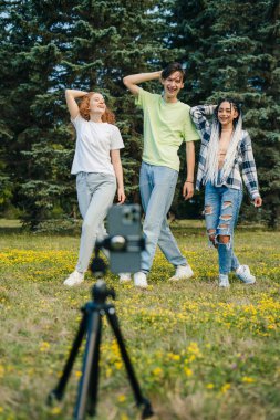 A boy and two girls of digital generation alpha cheering and dancing in front of a camera phone, while spending their weekend in the city park. Youth lifestyle