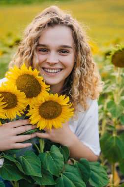 Portrait of a curly girl enjoying nature posing with a huge bunch of sunflowers in a summer evening. Toothy smile. Nature landscape. Summer nature. Harmony