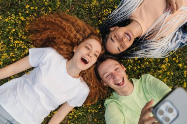 Top view of three smiling friends laying on grass holding a stick with mobile phone taking a selfie for social media. Outdoor recreation. Gen-z.
