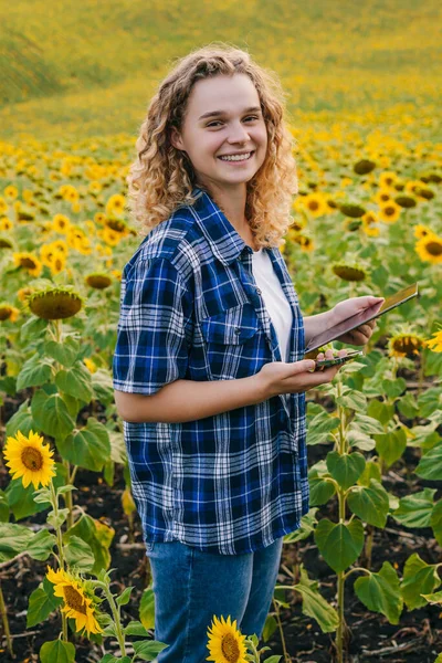 Portrait of a smiling curly girl working in sunflower field doing the analysis of growth of plant culture. Smart farming and precision agriculture. Sunflower
