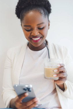 Beautiful Afro American woman drinking cold beverage and using mobile phone at cafe, smiling while resting in cafe. Different skin ethnicity female on remote