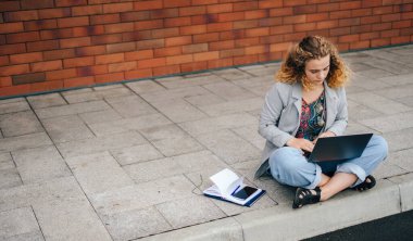 Curly-haired student woman learning a language over the Internet sitting on asphalt pavement using laptop. Looking at screen. Enjoying freelance remote job or