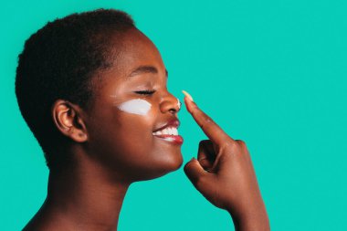 Profile view of african woman touching nose with index finger to apply cream isolated on sea green background with free space for text. Facial mask for skin