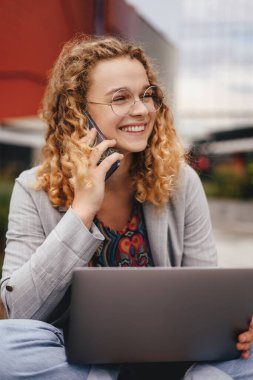 Smiling curly-haired woman student talking on cell phone while sitting with net-book in park during recreation time. Happiness and Education learning concept
