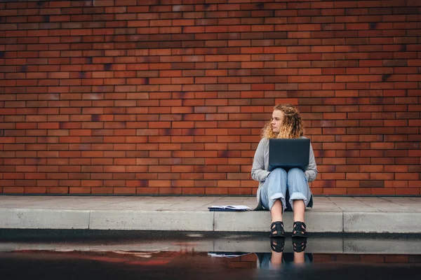Caucasian girl having training course via notebook device, sitting outdoors in city street on the background of a brick wall with free space for text