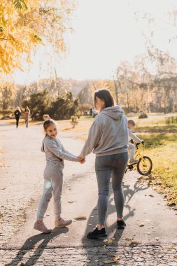 Back view of a mother spending time with her daughter walking in the park in the morning, the girl turning her head and smiling at the camera. Summer vacation