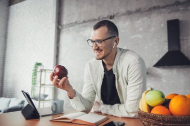 A caucasian man eating an apple looking into the tablet at the table early in the morning in a home kitchen. Business people communication. Kitchen interior