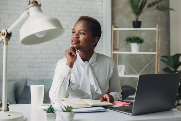 African american businesswoman looking away thoughtfully while working remotely with a laptop. Smiling, looking away, thinking over good news. Inspiration, idea