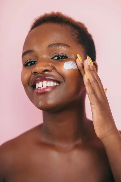 Portrait dark skinned woman with curly hair and toothy smile applying face cream on cheeks, isolated on pink background. Facial beauty. Beauty portrait. Beauty