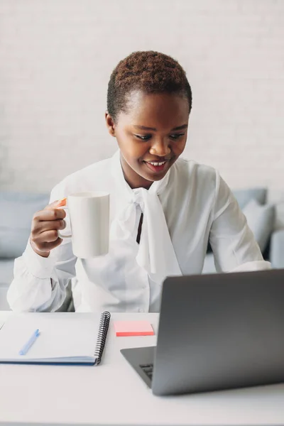 Portrait of a young african businesswoman sitting in office working on laptop computer while drinking a hot coffee. Cup of tea or coffee, papers and pens