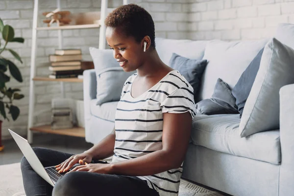 Young happy afro american woman, sitting on floor enjoying working online on laptop at home. Browsing internet, spending time at home in living room. Student