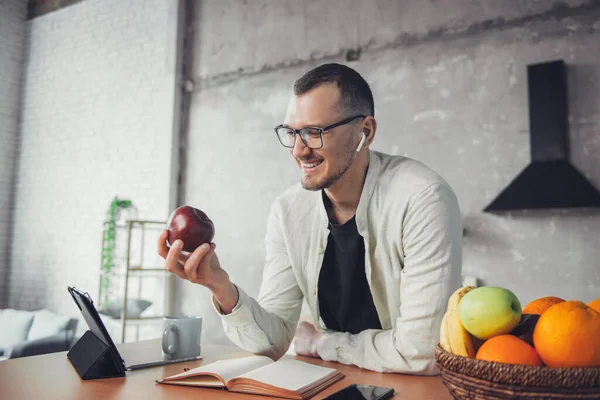 A caucasian man eating an apple looking into the tablet at the table early in the morning in a home kitchen. Business people communication. Kitchen interior