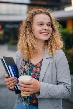 Close-up portrait of a pretty young student with curly-hair and jacket holding cup of coffee and laptop computer while standing in front of the university