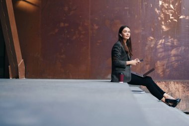 Young contemporary confident woman using gadgets, laptop, phone and wireless earphones, standing outside on the stairs at the entrance to the office building