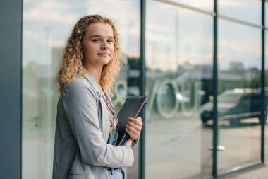Caucasian girl student holding notebook folders in her hands strolling near glass building looking at camera. Handsome woman waiting for someone. Breathing
