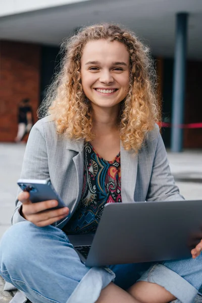 Smiling caucasian woman with mobile phone texting messages, working on laptop sitting outside. Communication, internet concept. Internet technology concept