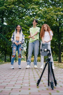 Modern trendy young people dancing in the summer in the park, recording a video with mobile phone. Gen Z group of friends using gadget and having fun outdoors.