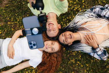 Happy three people lying down on green grass holding heads together looking up at the camera as posing for a selfie photo. New generation, gadgets easy usage in