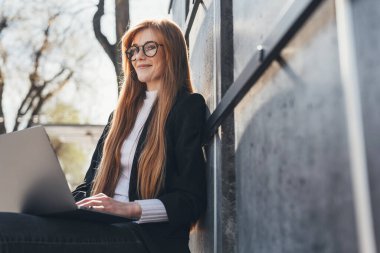 Beautiful businesswoman sitting on bench doing remote work outdoors using laptop. Browsing internet and searching for information. Freelancer working