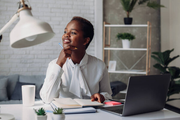 Attractive happy stylish African black woman working at laptop computer at office, dreaming about weekend vacation. Positive thinking person concept. Office