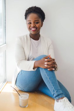 African woman in jacket sitting on window sill, relaxing in coffee shop with a glass of iced coffee. Relaxation or weekend away from home.