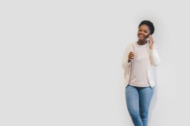Beautiful afro american woman using mobile phone in the coffee shop leaning against the white wall with copy space. Drinking coffee at the workplace. Wireless