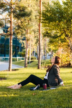 Side view of a woman relaxing on the grass field of the park in the morning on a sunny day, having break at work. Beautiful young girl. Summer season.