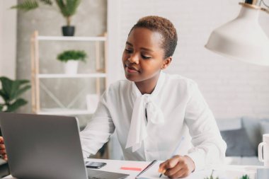 Photo of cheerful mixed-race woman in white shirt working with laptop while sitting at table in office. Teamwork, business meeting. Freelancers working. Student