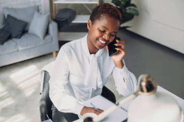 Busy young afro woman talking on cellphone with client and taking notes, sitting at workplace in modern office. Education internet technology. Marketing