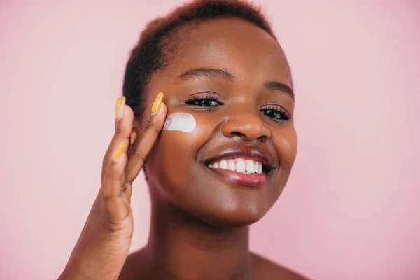 Beautiful black african model with flawless skin putting cream treatment before makeup cosmetic routine at home, isolated over pink background. Happy young