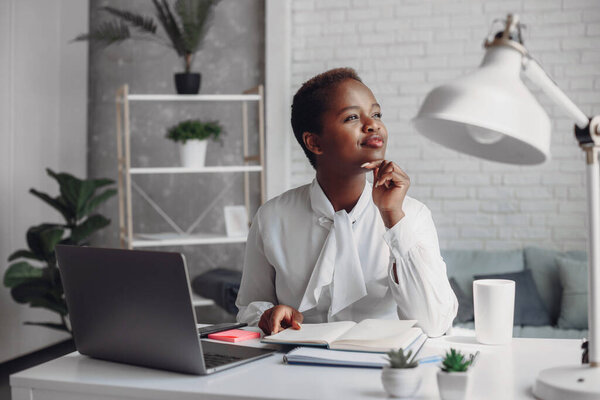 Pensive mixed race female entrepreneur sitting in home office, looking away at window, thinking of future vision. African business woman having break in work