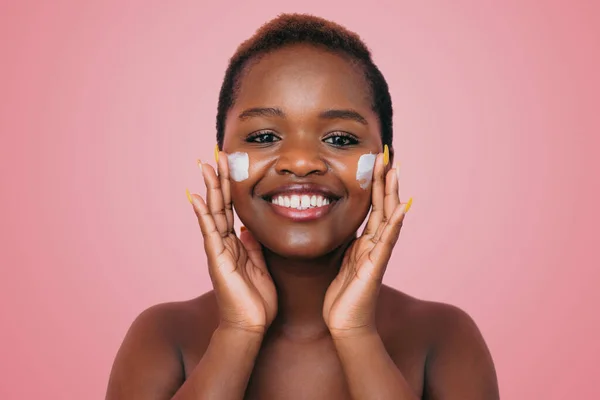 Close up shot of positive dark skinned woman applying moisturizing cream on her face isolated on pink background. Skin care and beauty concept. Dermatology