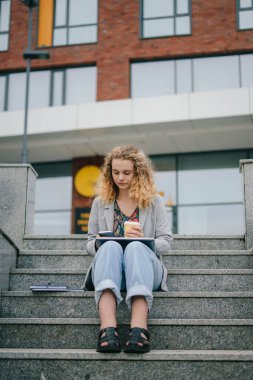 Modern beyaz kadın öğrenci dışarıda ders çalışıyor, bir fincan kahve servisi ve merdivenlerde oturan akıllı telefonları kullanarak internette geziniyor. Mesafe çalışması veya eğitim