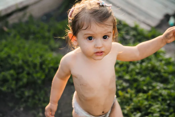 Little baby girl with dirty face and dirty body looking on camera, and pointing with his hand to something she found while in the garden. Baby wearing white