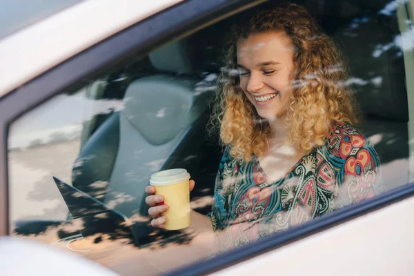 View from the car window smiling young woman, student girl checking e-mail on laptop, while drinking coffee sitting in the car while traveling. Sunset, concept