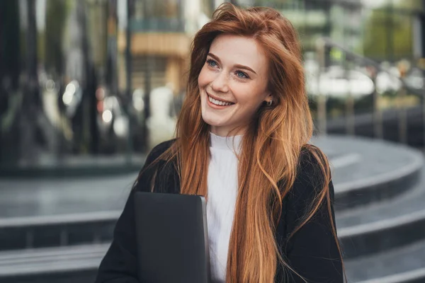 Young confident businesswoman in formalwear holding laptop in hands while leaving office building after hard working day. Job interview. Woman standing outdoor