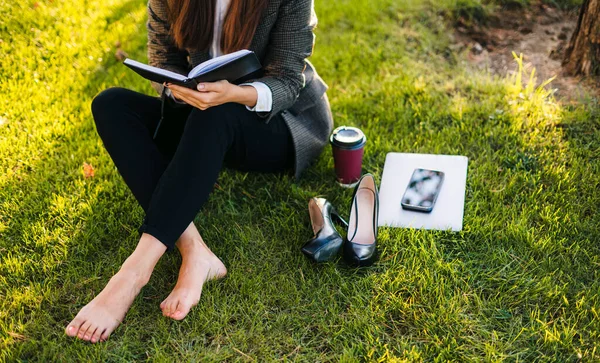 Cropped view of a beautiful caucasian brunette businesswoman sitting barefoot on the green grass reading a book in the city park. University girl reading books