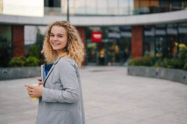 Ders kitaplarını tutan başarılı öğrencinin portresi gülümseyerek başını modern üniversite binasının önünde kameraya çeviriyor. Öğrencilik