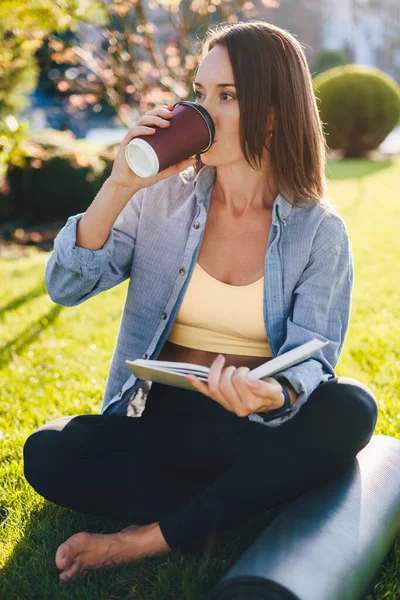 Portrait of smiling caucasian woman drinking morning coffee, reading text book sitting near yoga mat on grass field. Outdoor education lifestyle concept