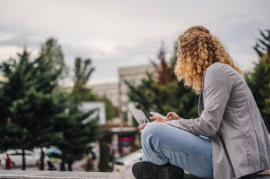 Açık havada ders çalışan gri ceketli genç bir üniversite öğrencisinin yan görüntüsü. Üniversitenin dışında laptopunu kullanıyor. Metin için boş alan. Uzak çalışma
