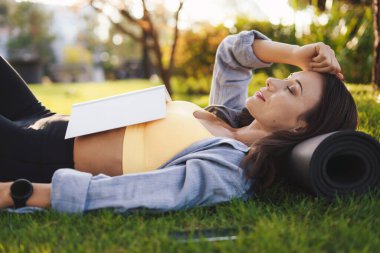 Sportif bir kadın yorucu bir sabah antrenmanından sonra, göğsünde kitap tutarak parkta yatıyor. Ev karantinası. Yoga yapıyor ve kitap okuyor.