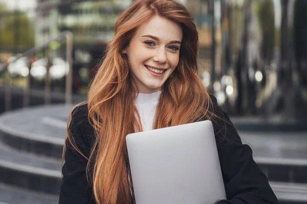 Happy attractive female freelancer with a laptop standing on city street staring at camera. Network communication, computer work, social distance. Freelance